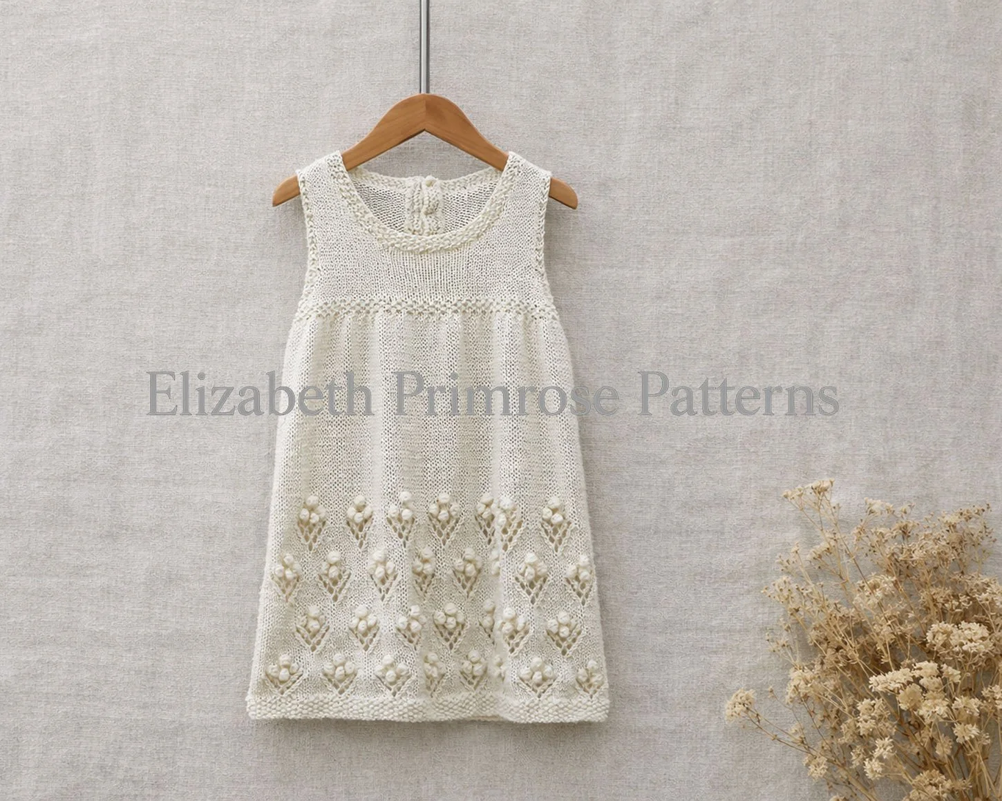 Cream knitted baby dress featuring a textured bobble flower skirt, displayed on a hanger against a beige backdrop with a small bunch of dried flowers.
