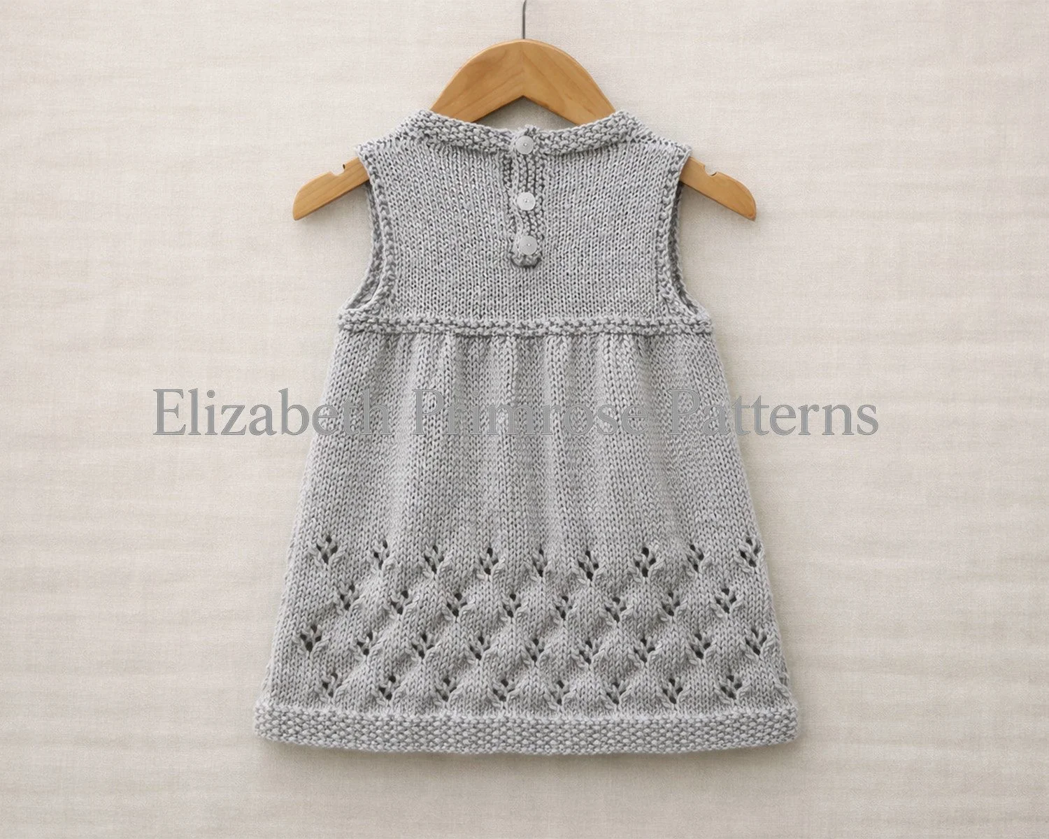 Back view of a grey hand knitted sleeveless baby dress, hanging on a wooden hanger over an uncreased light beige linen background.