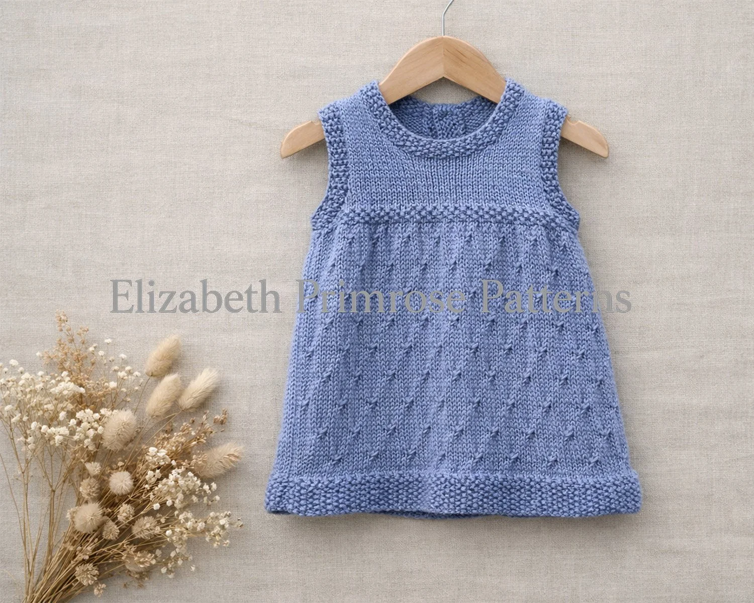 Grey knitted baby dress on a wooden hanger against a beige linen background, with delicate diamond texture and dried flowers arranged to the side.