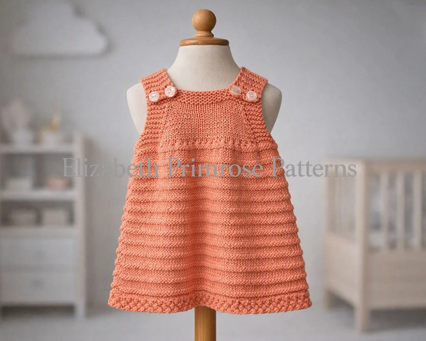 Hand‑knitted baby pinafore dress on a fabric mannequin with textured ridges and bobble hem in a softly lit nursery setting.