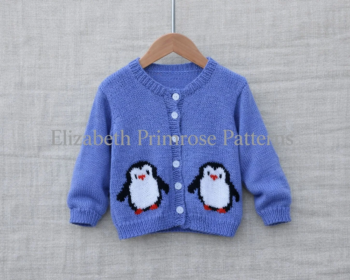 Hand-knitted blue children’s cardigan with penguin motifs, displayed on a wooden hanger against a soft beige linen background.