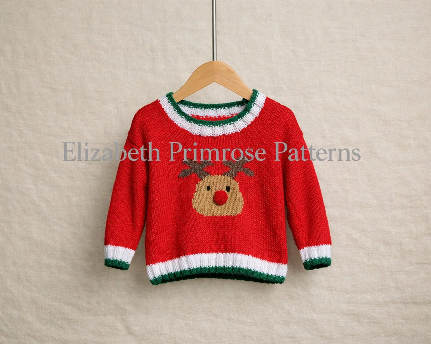 Red knitted children’s jumper on a wooden hanger with a reindeer motif and striped trims against a soft beige linen background.