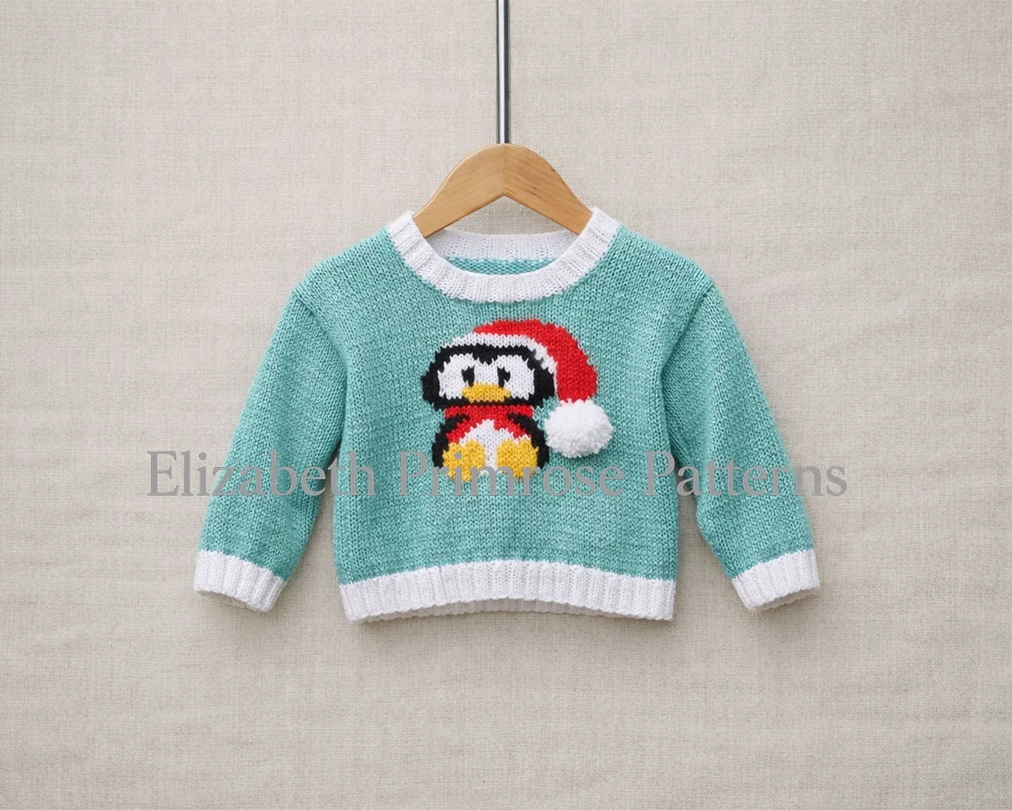 Light turquoise children’s jumper with white ribbing and a festive penguin motif, displayed on a wooden hanger against a soft beige fabric background.