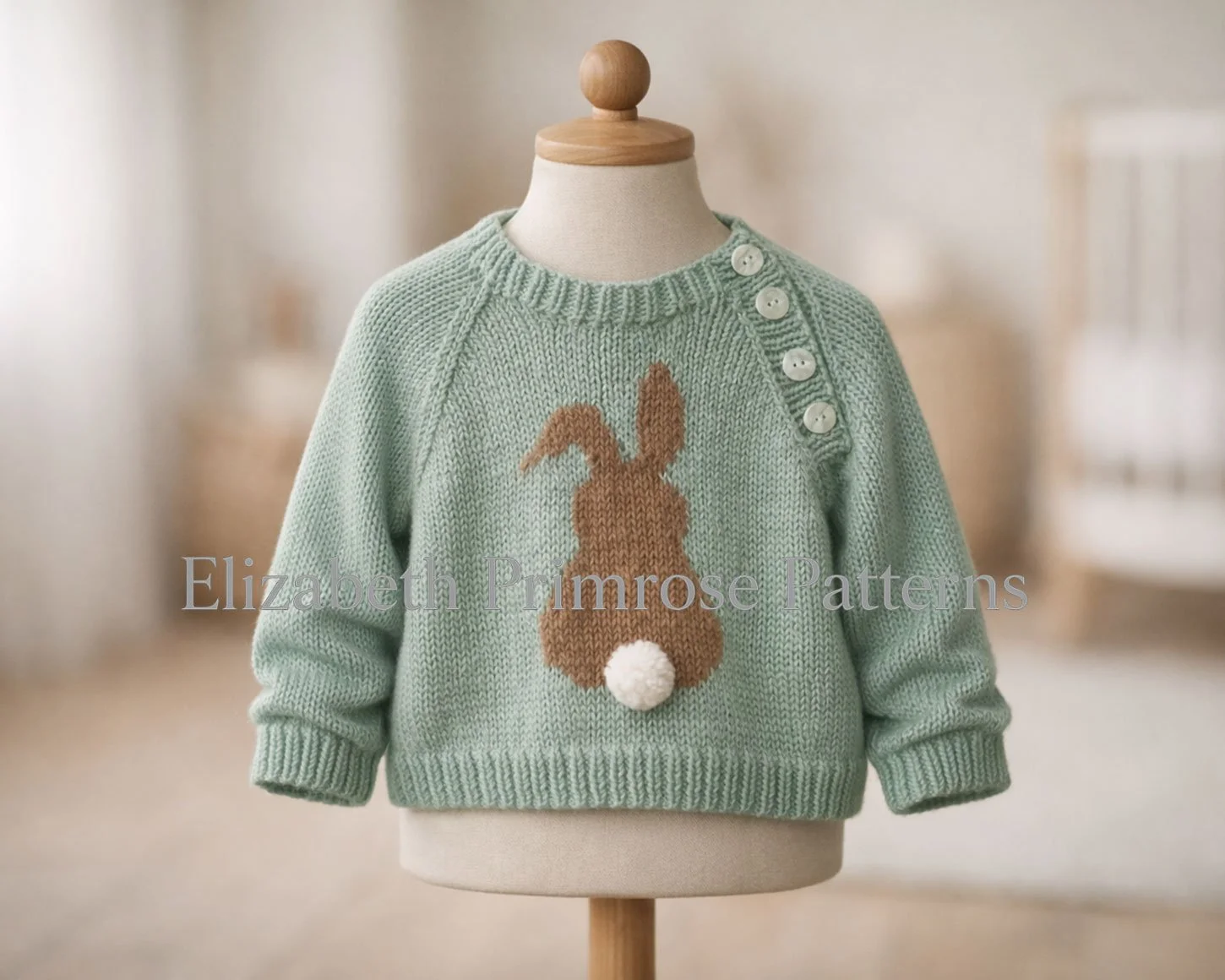 Sage green knitted baby sweater featuring bunny design and pearly shoulder buttons, shown on child mannequin with relaxed sleeves in warm, blurred nursery setting.