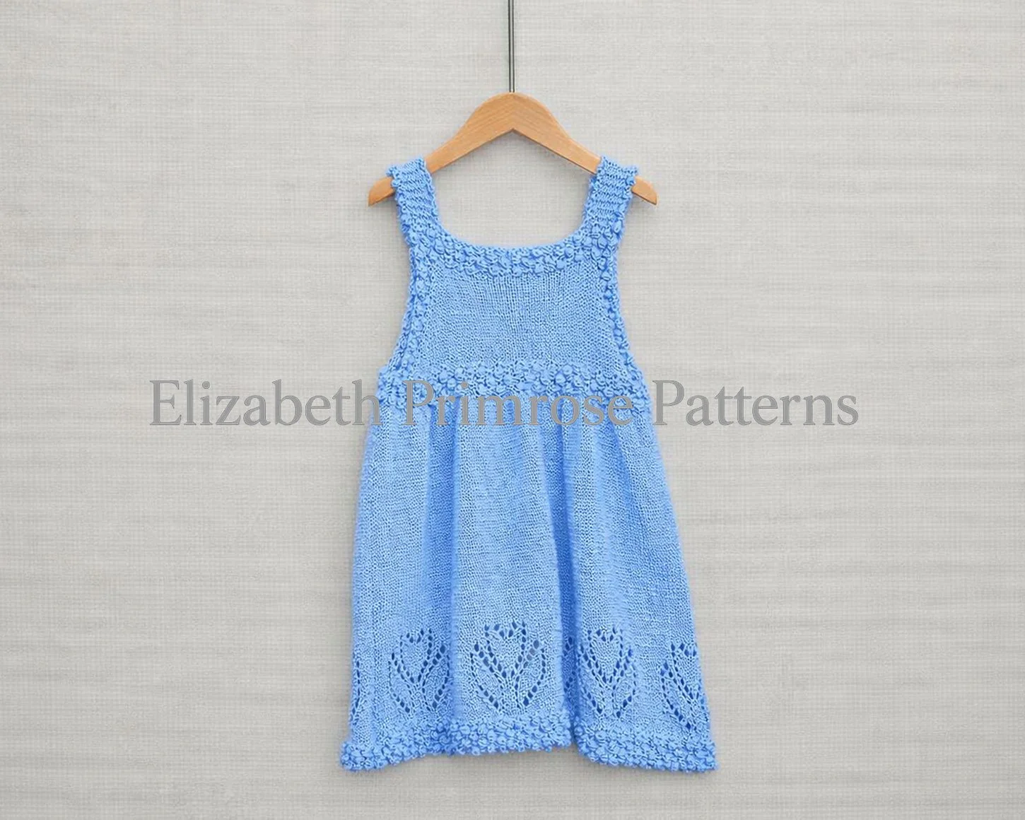 Back view of light blue knitted child’s dress with textured straps and floral motifs, displayed on hanger with neutral backdrop and dried flowers.