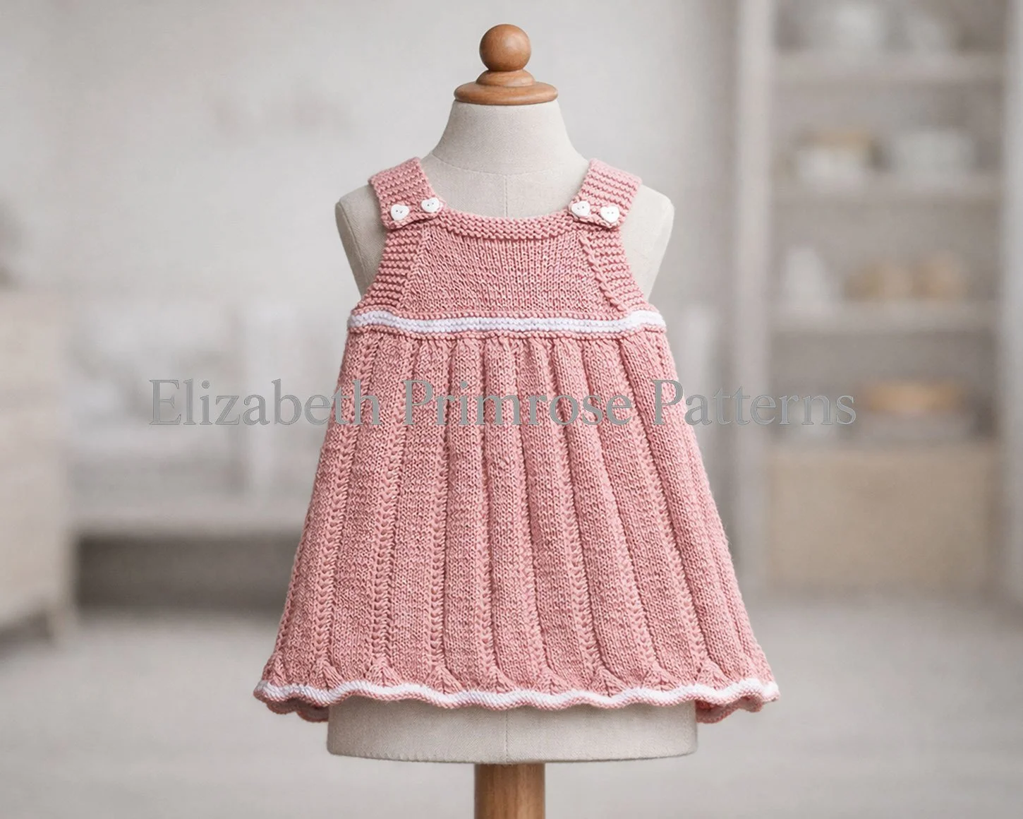 Hand knitted pink baby pinafore dress on a child’s mannequin, showing crisp stitch detail and wavy hem in a soft, minimalist room.