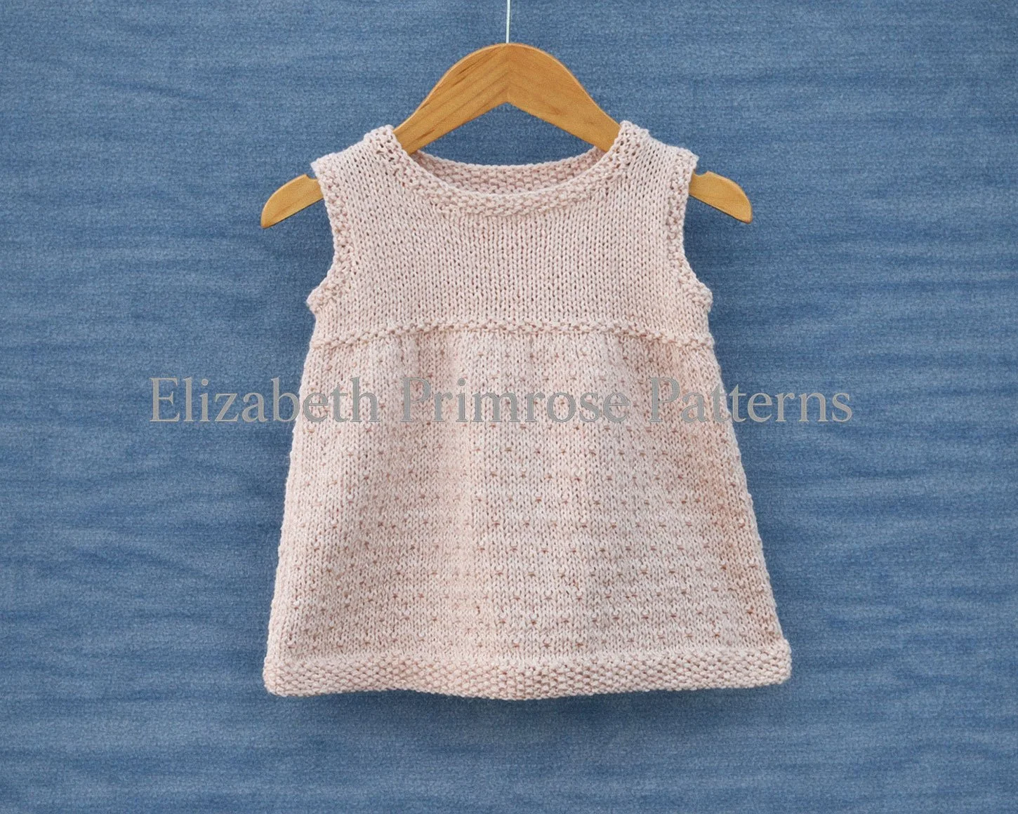 Sleeveless cream dress with dotty skirt pattern and moss stitch edgings on a hanger and blue background.
