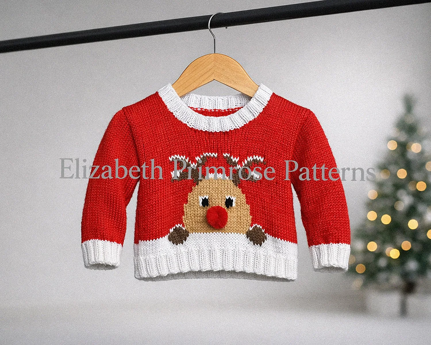 Hand knitted red festive sweater with white trims and a Rudolph reindeer motif, displayed on a wooden hanger against a blurred Christmas tree background with warm lights.