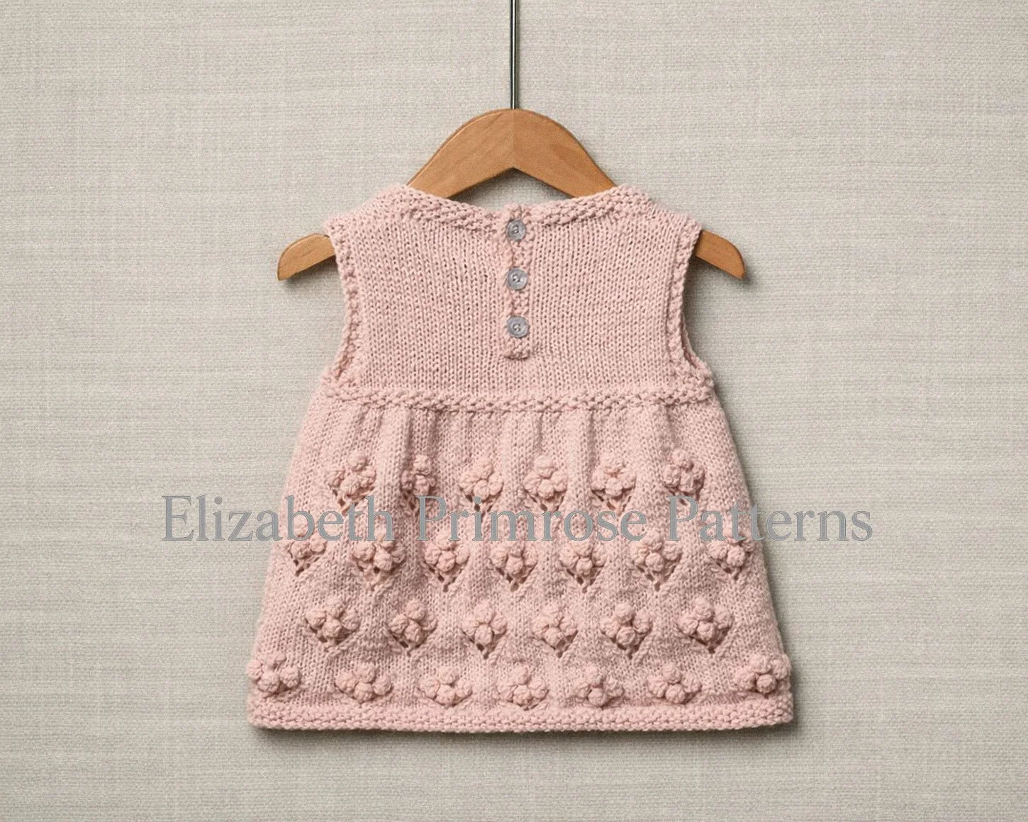 Back view of pink hand knitted sleeveless baby dress with bobble flower skirt, 3 button fastening, hanging on a wooden hanger and shown against a neutral fabric background.