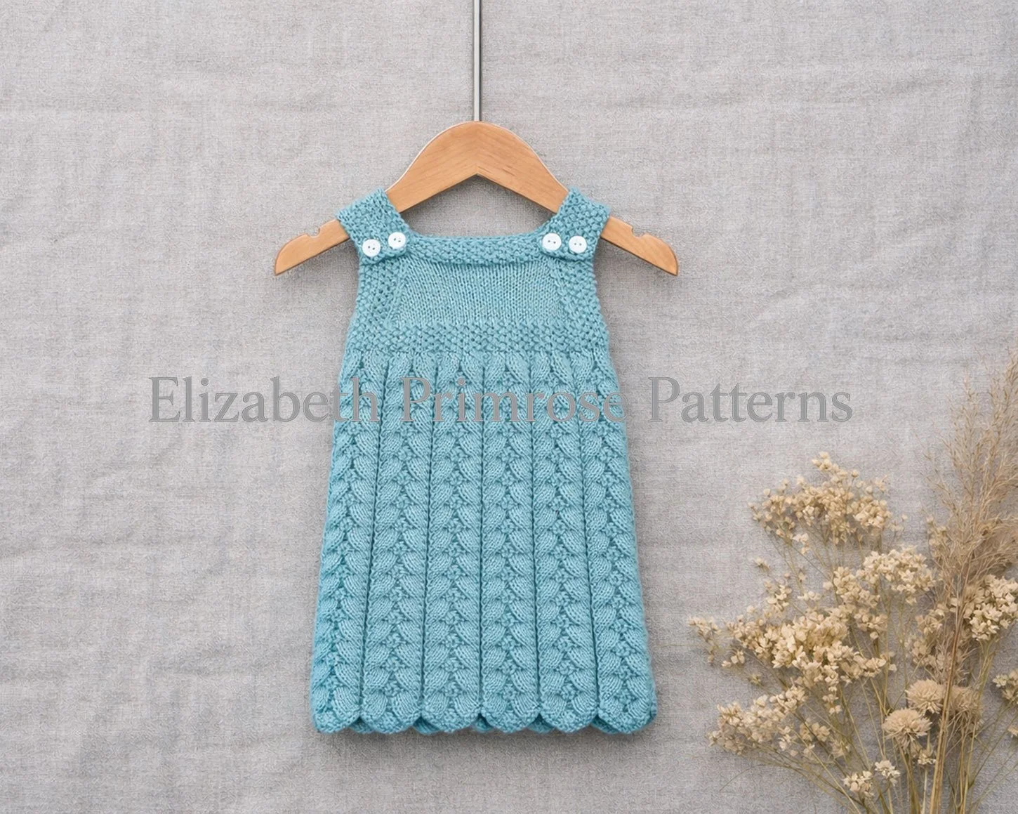 Hand knitted turquoise baby dress on a wooden hanger with textured cables, scalloped hem, white buttoned straps, and dried flowers beside it against a soft fabric backdrop.