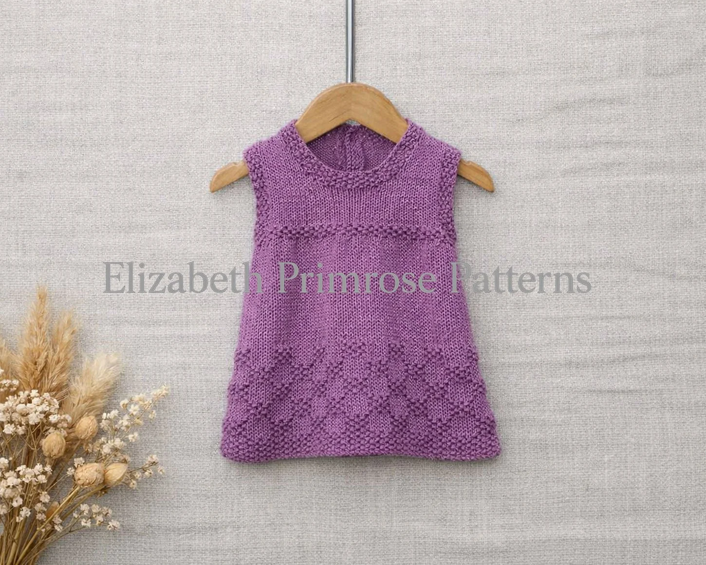 A hand‑knitted, sleeveless purple children’s dress on a wooden hanger, shown against textured beige linen with dried flowers in the corner.