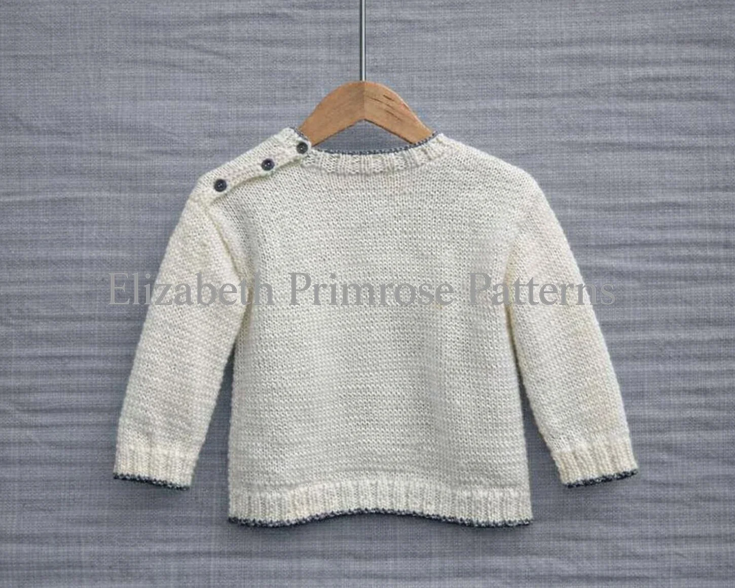 Back view of a cream knitted children’s jumper on a wooden hanger, with button opening to shoulder, shown against a sharp grey linen background.