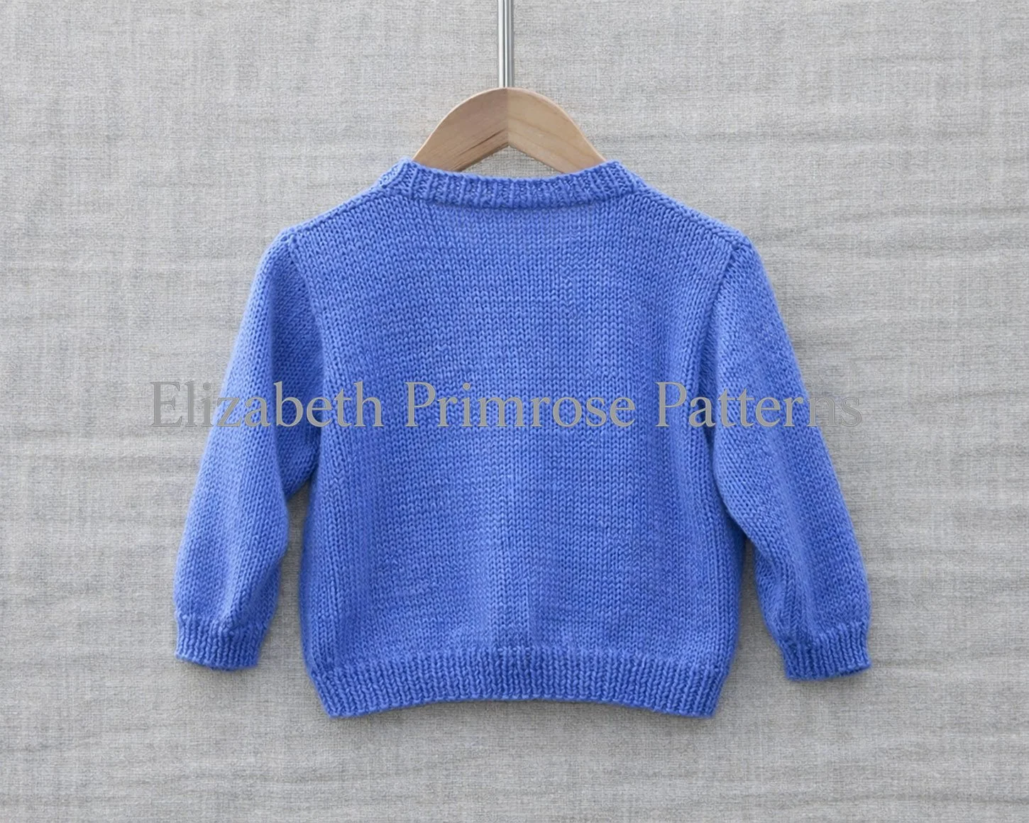 Back view of hand-knitted blue children’s cardigan, displayed on a wooden hanger against a soft beige linen background.