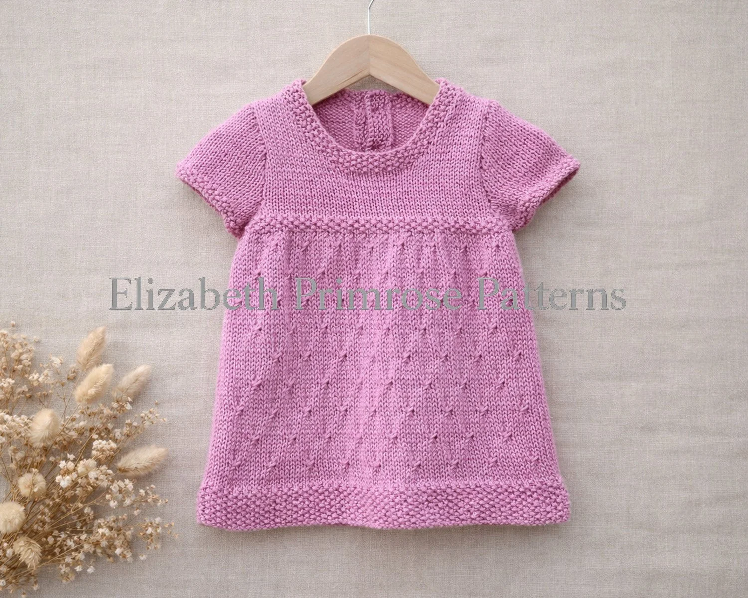 Pink knitted, short sleeved baby dress on a wooden hanger against a beige linen background, with delicate diamond texture and dried flowers arranged to the side.