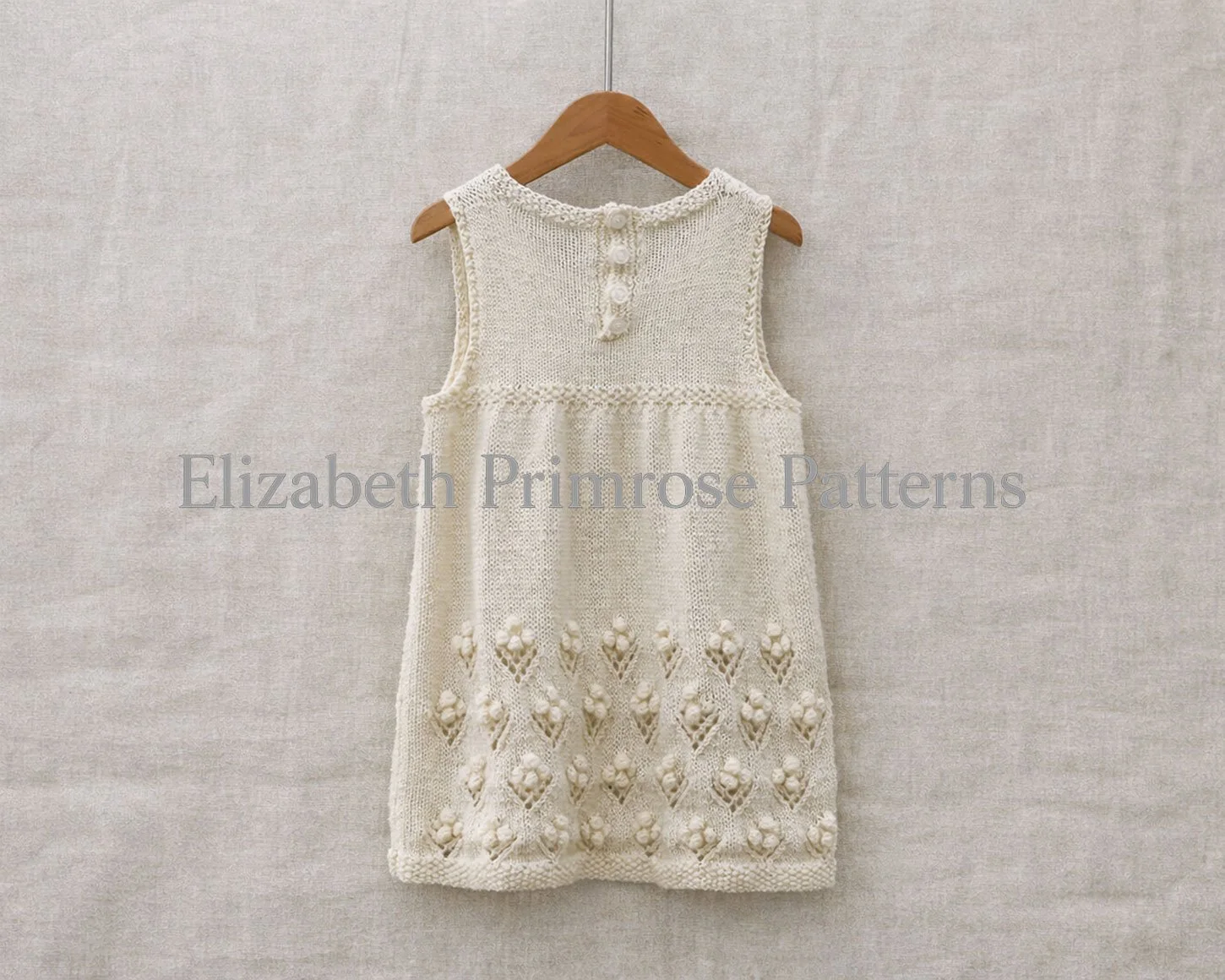 Back view of cream hand knitted sleeveless baby dress with bobble flower skirt, 4 button fastening, hanging on a wooden hanger and shown against a neutral fabric background.