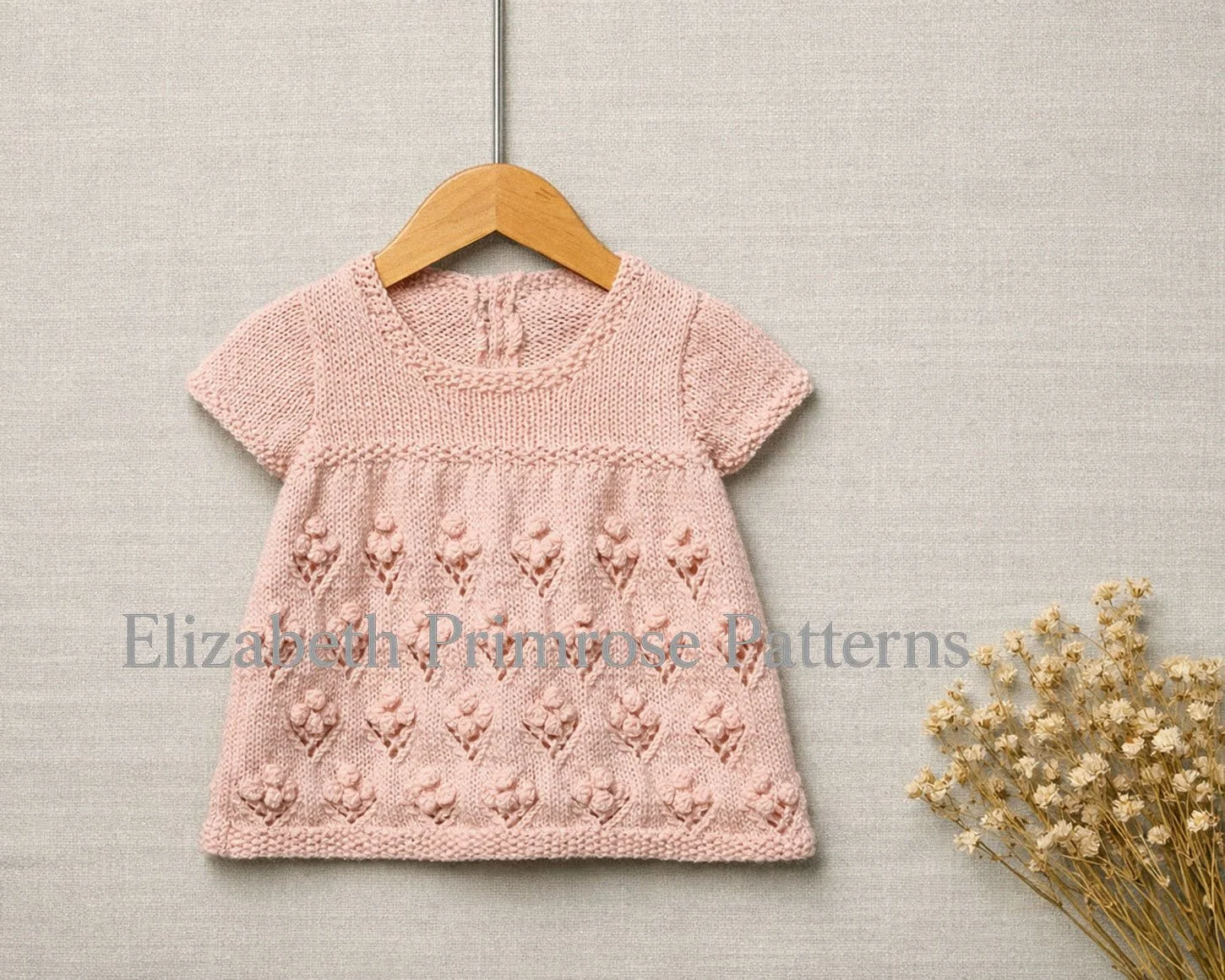 Pink hand knitted short-sleeved children’s dress with bobble flower skirt hanging on a wooden hanger, shown against a soft neutral fabric background with dried flowers.