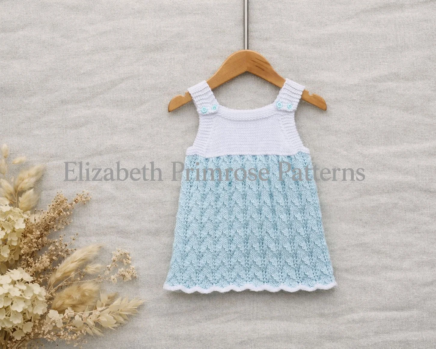 Knitted baby dress on wooden hanger with wave patterned blue skirt and white bodice, dried flowers arranged softly to the side.