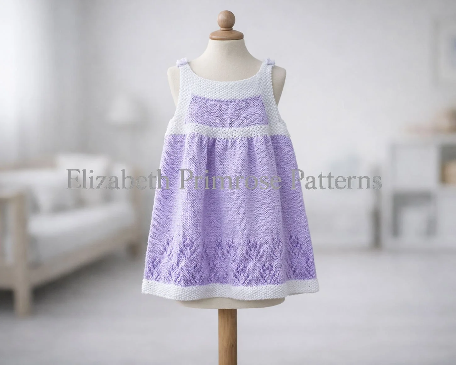 Lavender and white knitted pinafore dress with lace flower pattern shown on a child’s mannequin against a softly lit Scandinavian‑style background.
