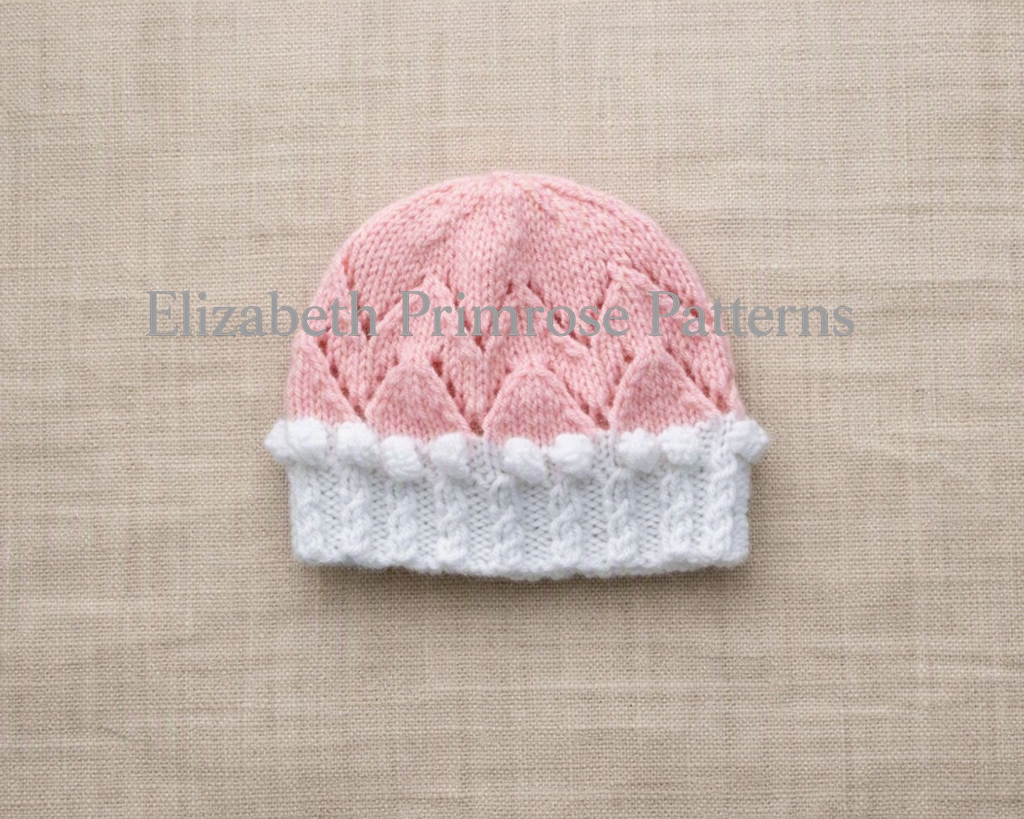 Valentina hand-knitted pink doll hat with white lace trim and bobbles, laid flat on a textured neutral background.