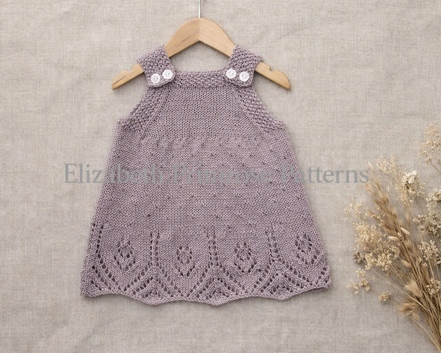 Lavender knitted pinafore dress on a wooden hanger against a soft fabric backdrop, featuring textured dot pattern, lace hem detail and buttoned straps with dried flowers beside it.