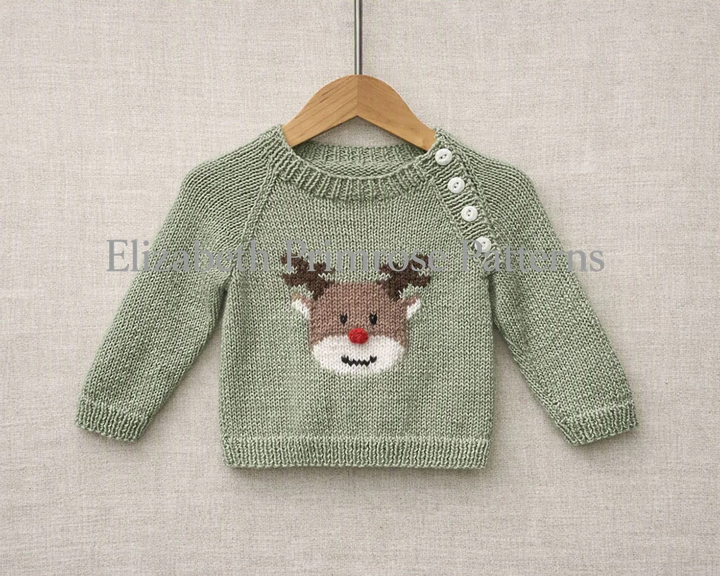 Light green raglan baby sweater with reindeer motif and buttoned shoulder, displayed on wooden hanger against beige fabric background. Knitting pattern by Elizabeth Primrose Patterns.