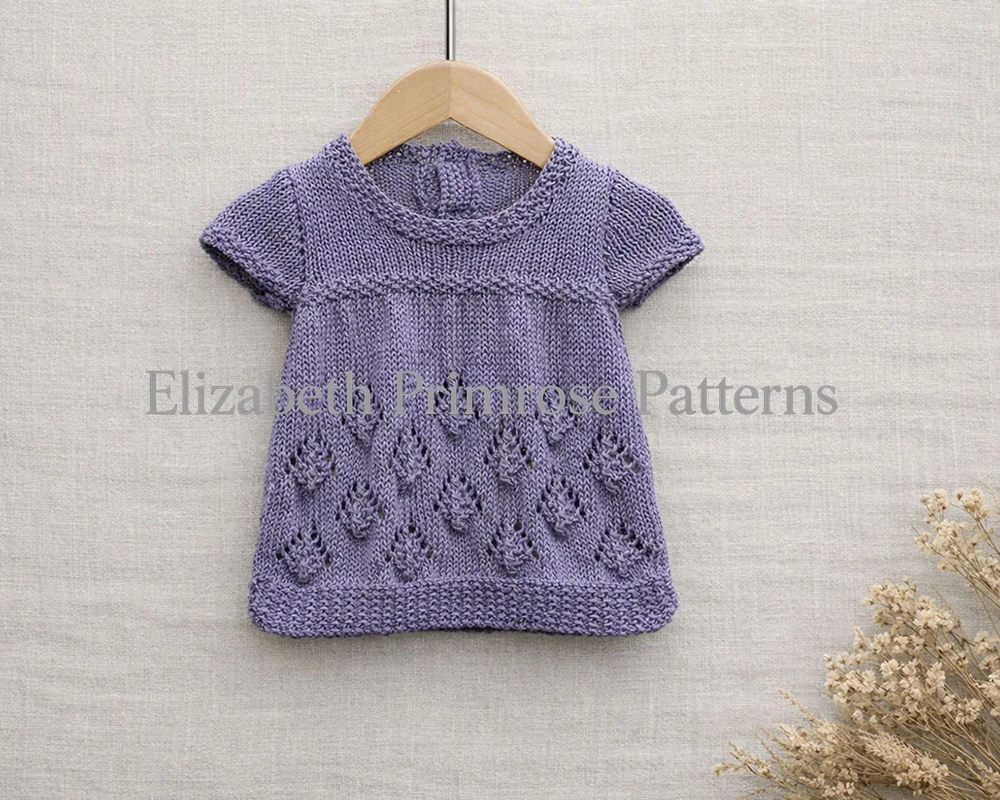 Lavender knitted baby dress with pinecone pattern skirt hanging from a wooden hanger against a light beige linen backdrop with dried flowers.