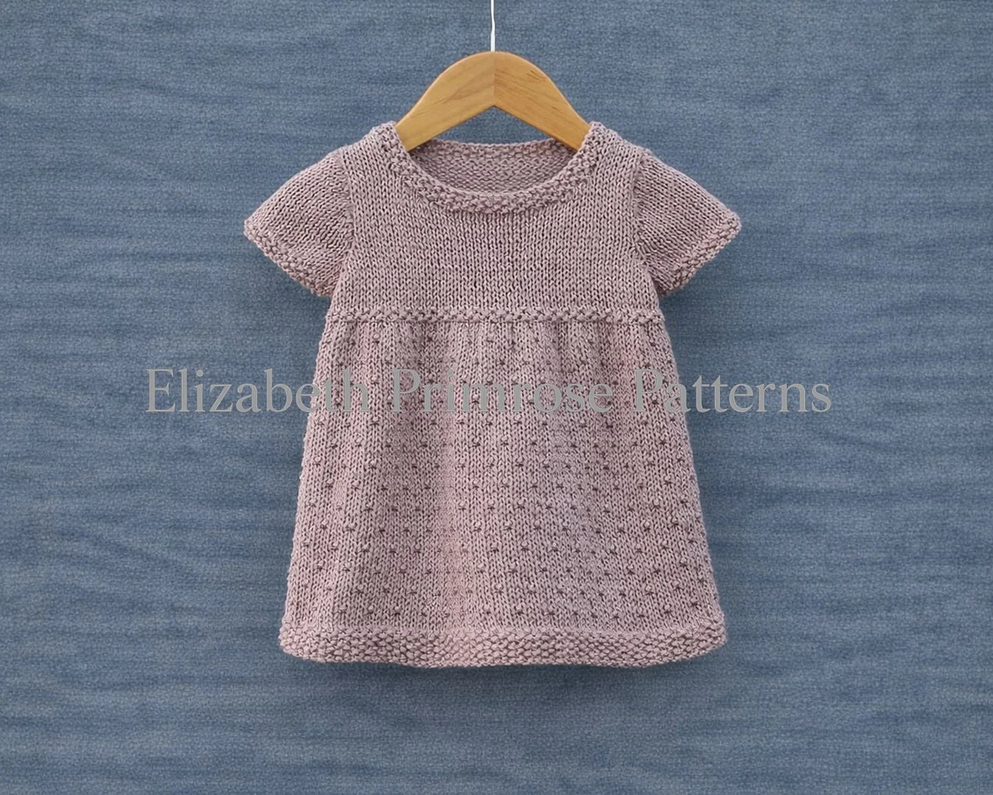 Short sleeved mauve dress featuring a textured skirt and moss stitch edgings on a hanger and blue background.