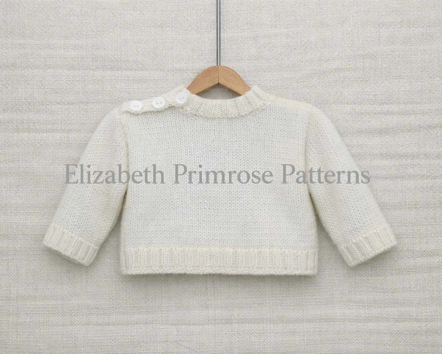 Back view of cream knitted baby jumper with buttoned shoulder, hanging on a wooden hanger against a beige fabric backdrop.
