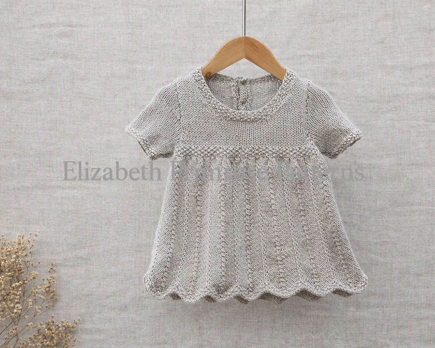 Light grey hand‑knitted children’s dress with chevron skirt and scalloped hem hanging on a wooden hanger against a neutral fabric backdrop.
