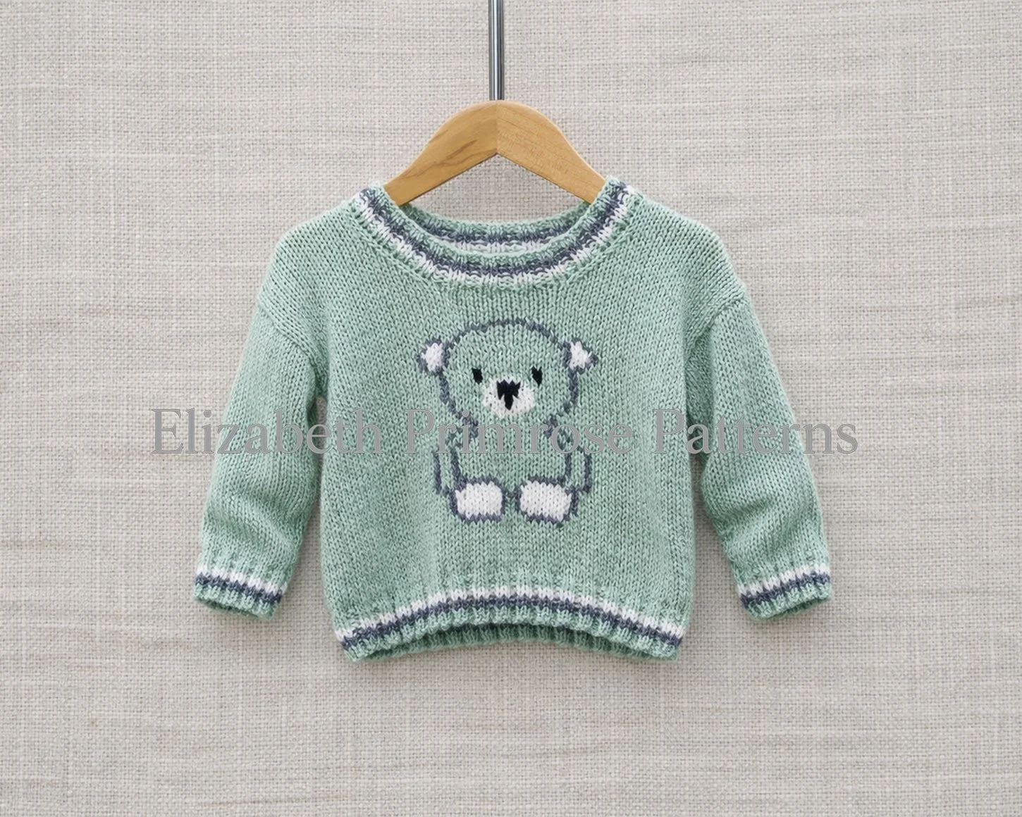 Light green hand-knitted baby jumper with bear motif, hanging on wooden hanger against beige fabric background, labelled Elizabeth Primrose Patterns.