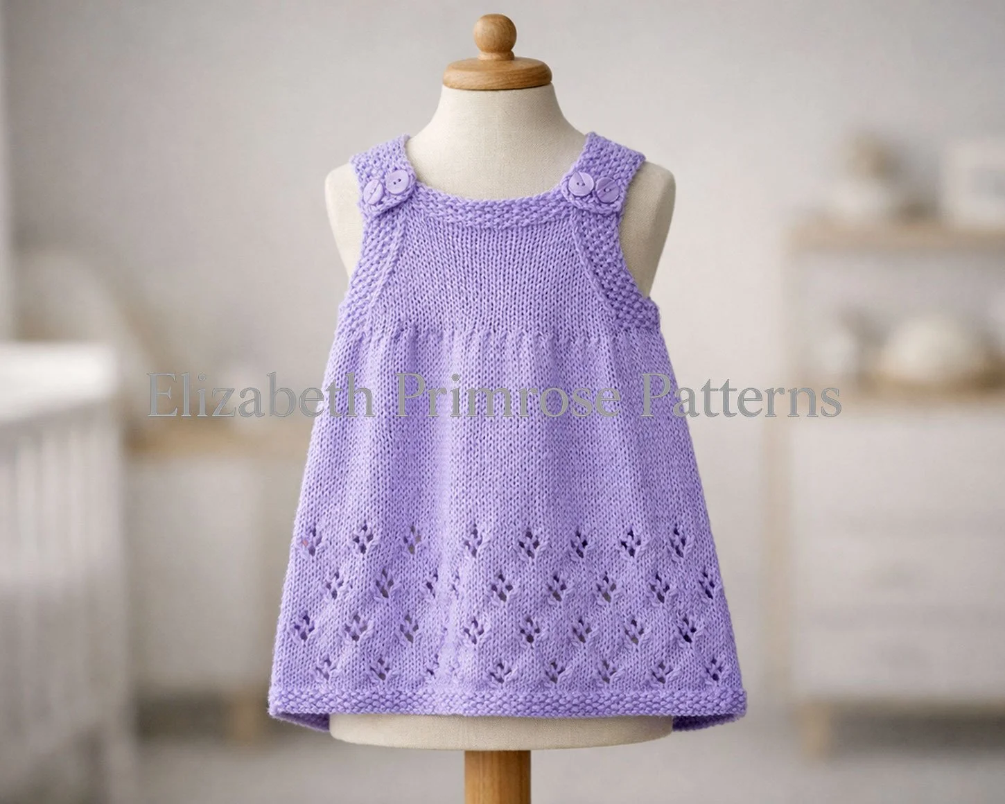 Lilac hand knitted children’s pinafore dress on a mannequin, featuring buttoned straps, moss stitch edges, and a delicate lacy snowflake skirt pattern in soft daylight.
