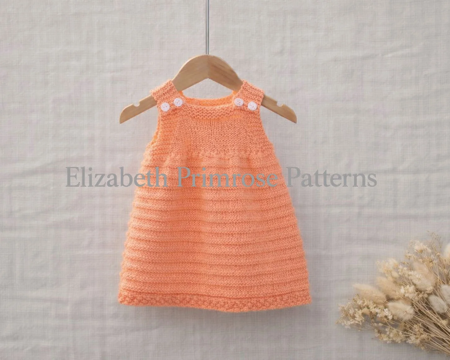 Peach knitted baby dress with buttoned straps hanging on a wooden hanger against a linen background with dried flowers.