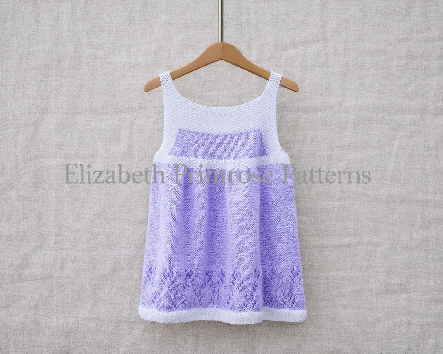Back view of hand‑knitted children’s dress in lavender and white displayed on a hanger on a linen background.