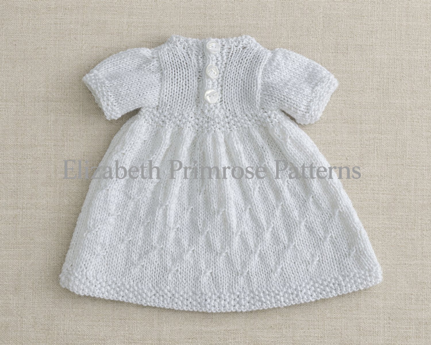 Back view of white knitted doll dress laid flat on textured fabric, featuring a patterned skirt and three small buttons.