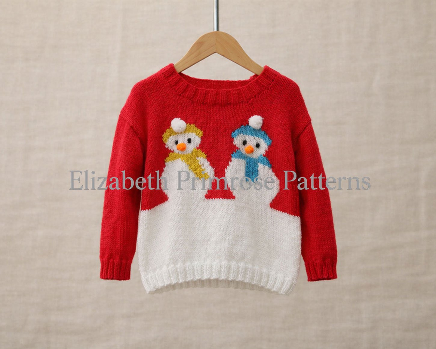 Red and white children’s knitted jumper with two snowmen in yellow and blue scarves, hanging on a wooden hanger against a soft beige background.