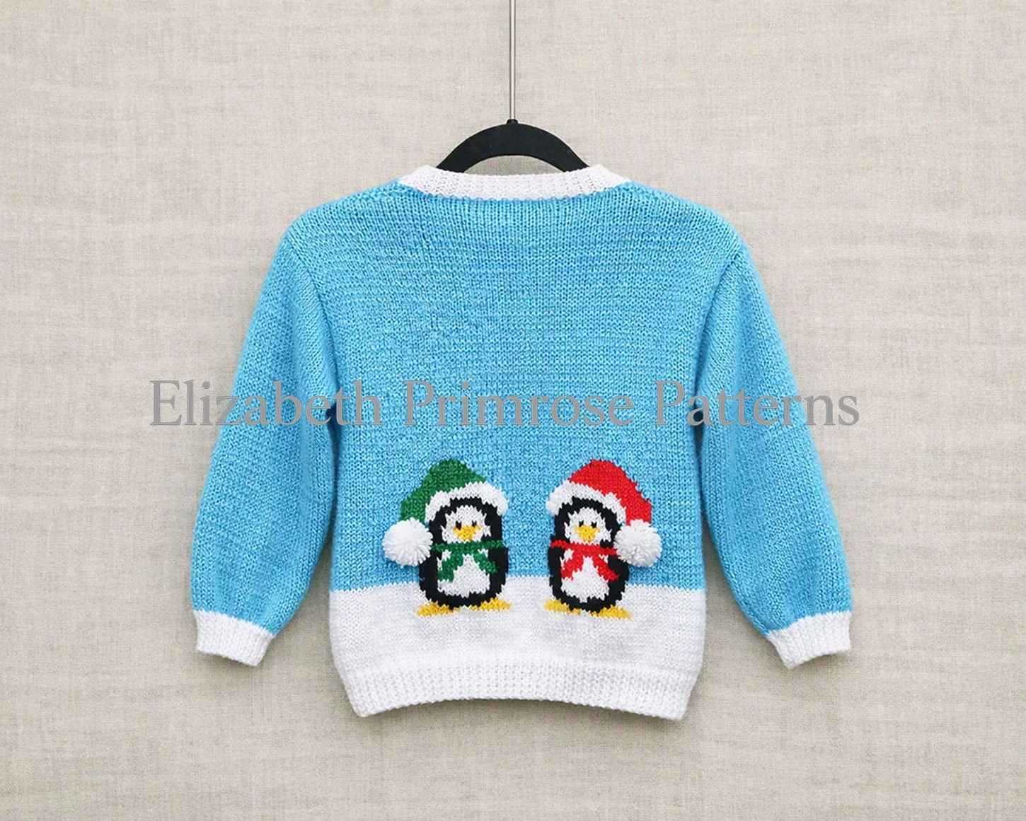 Back of a blue children’s jumper with two penguins in winter hats, hanging on a wooden hanger against a smooth beige linen background.