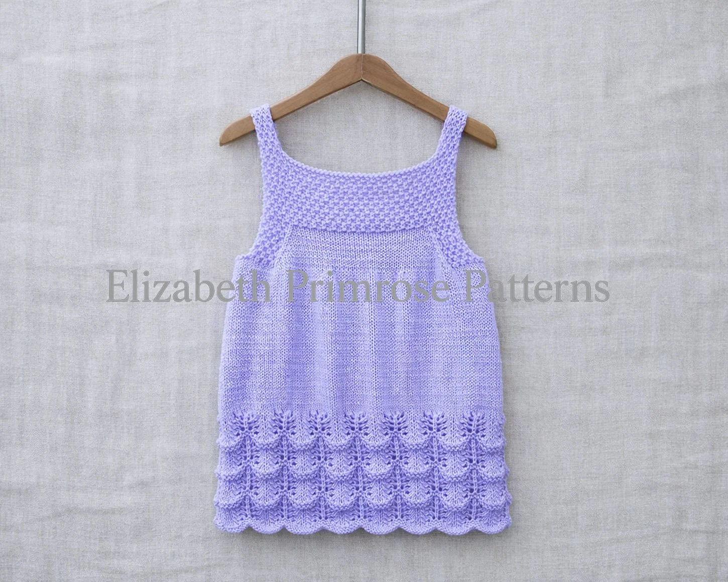 Back view of lilac hand knitted baby dress with scalloped lace hem hanging on a wooden hanger against a soft linen backdrop.