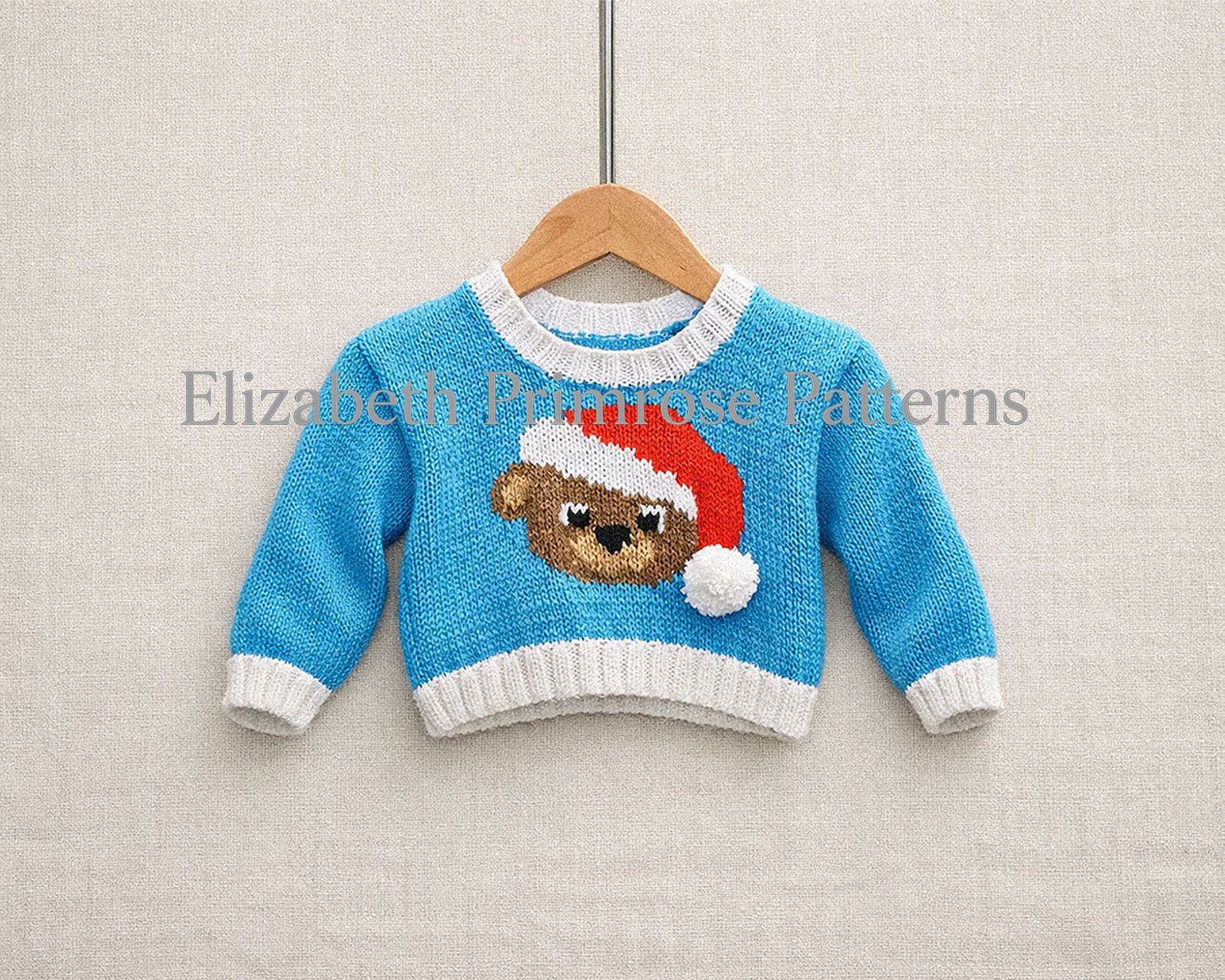Festive blue baby jumper with white trims and a teddy bear in a Santa hat, hanging neatly on a wooden hanger over a smooth beige fabric backdrop.