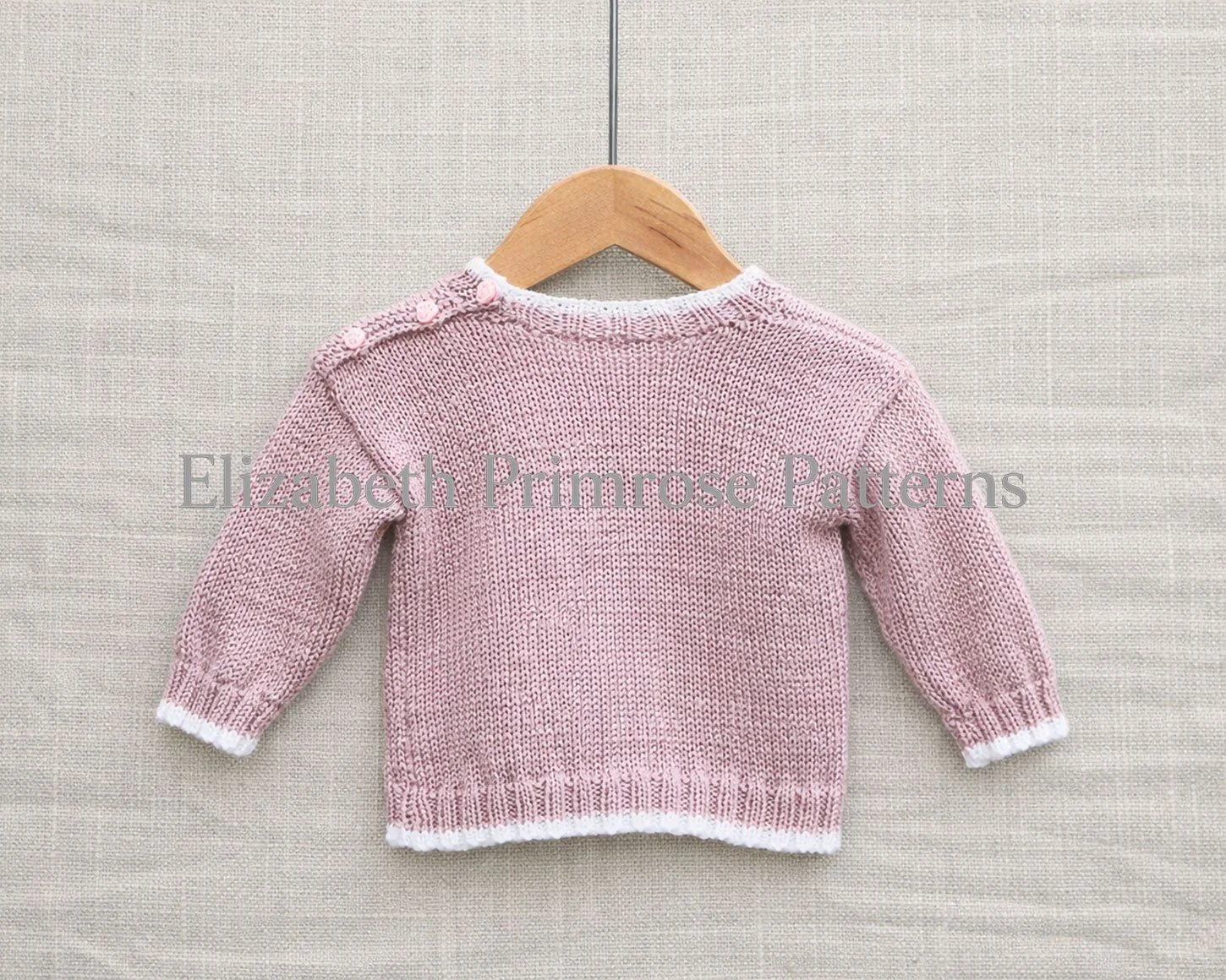 Back view of pink knitted baby sweater with relaxed sleeves and white ribbed edges on linen backdrop.