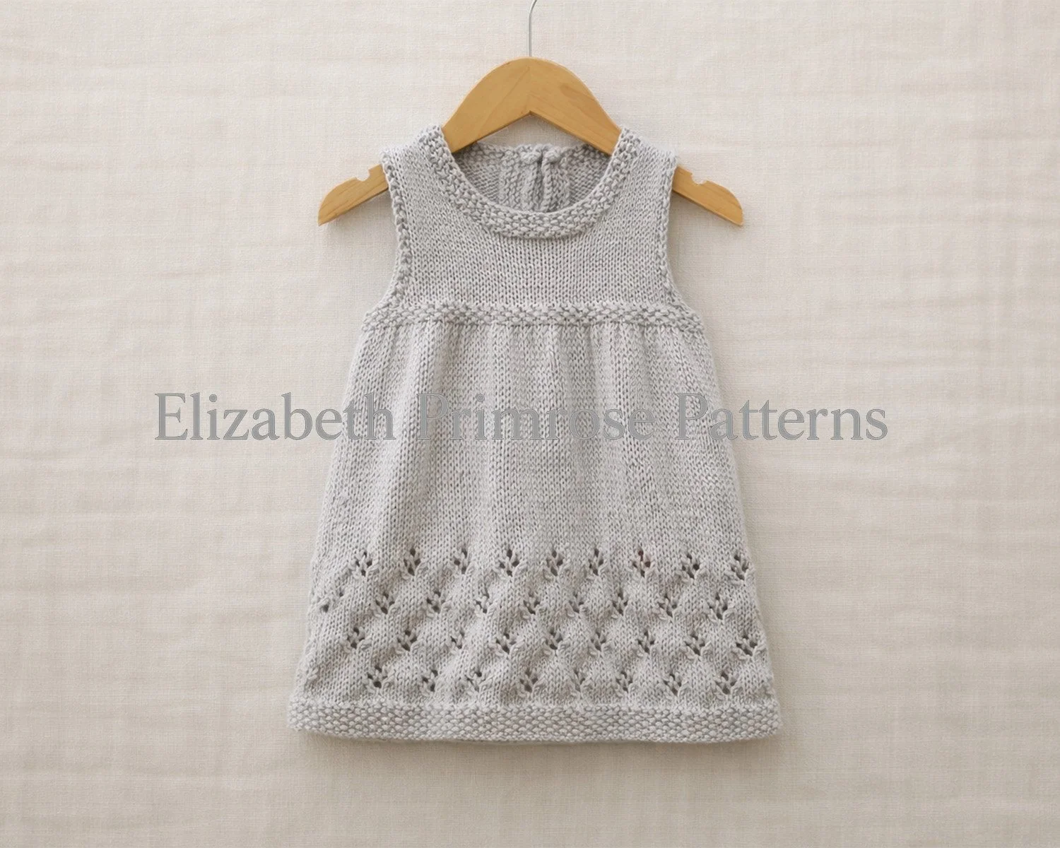 Light grey hand knitted sleeveless baby dress hanging on a wooden hanger against an uncreased light beige linen flatlay background.