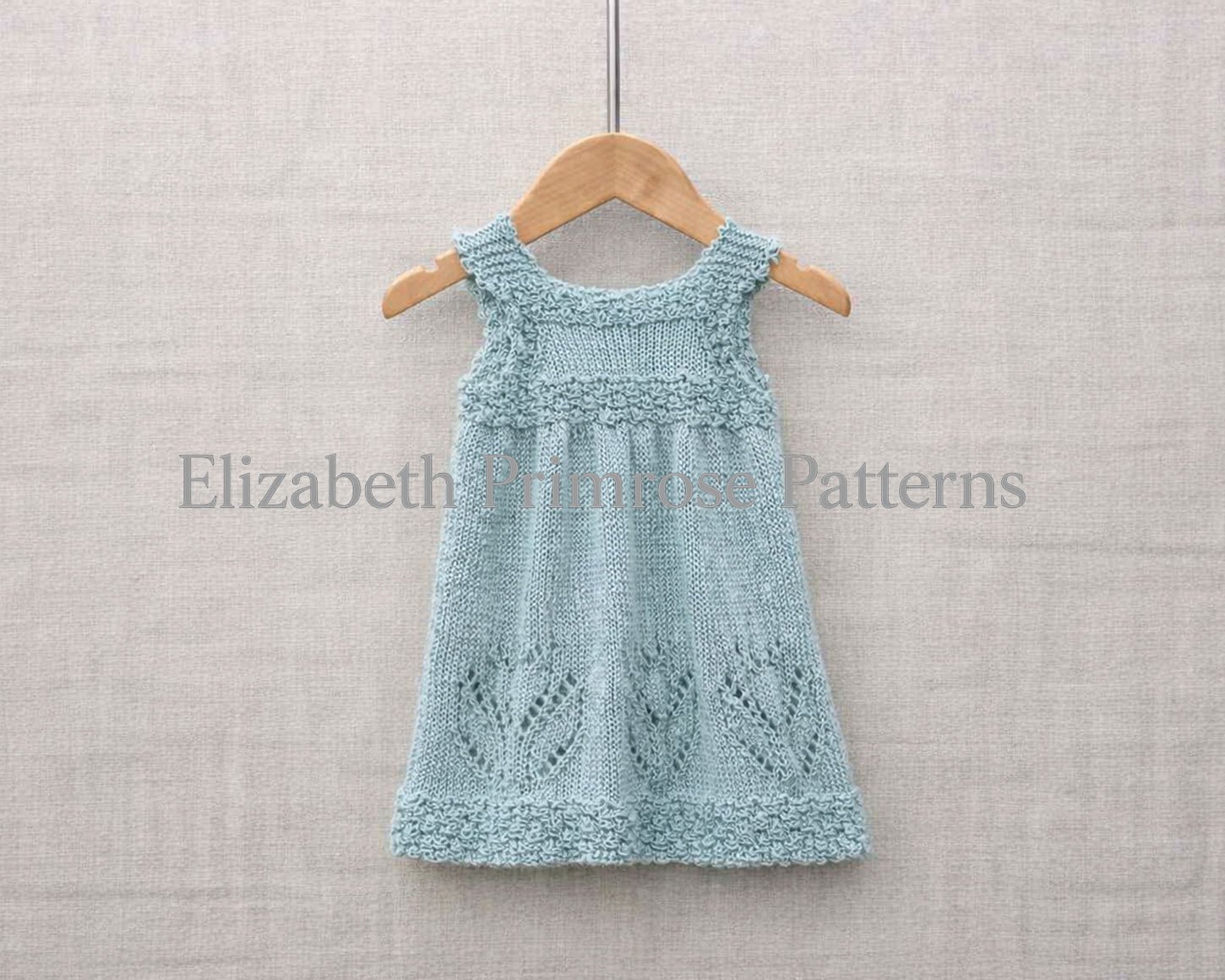 Back view of a pale blue knitted baby dress on a wooden hanger with crisp lace tulip motifs and bobble details against a neutral fabric background.