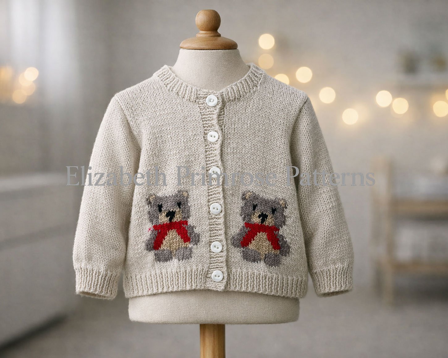 Cream knitted baby cardigan featuring bear designs and white buttons, hanging naturally on a mannequin with extended fabric torso and blurred fairy‑lit background.