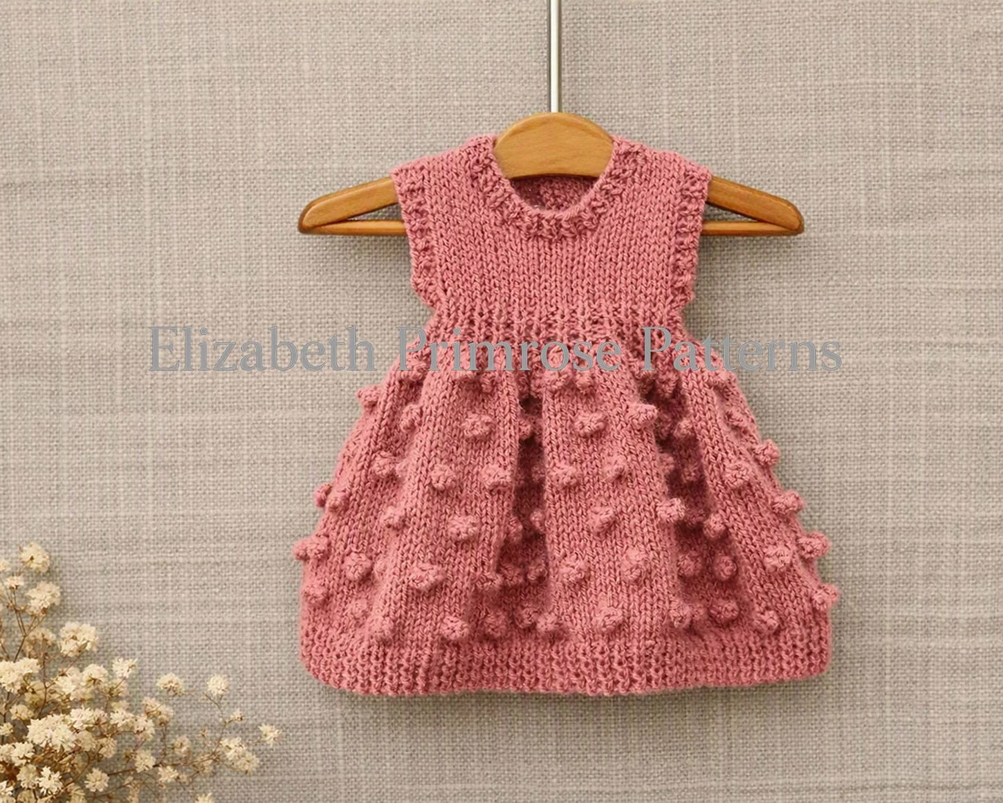 A hand-knitted pink doll dress hanging on a wooden hanger against a beige background, showcasing a sleeveless version with bobble skirt.