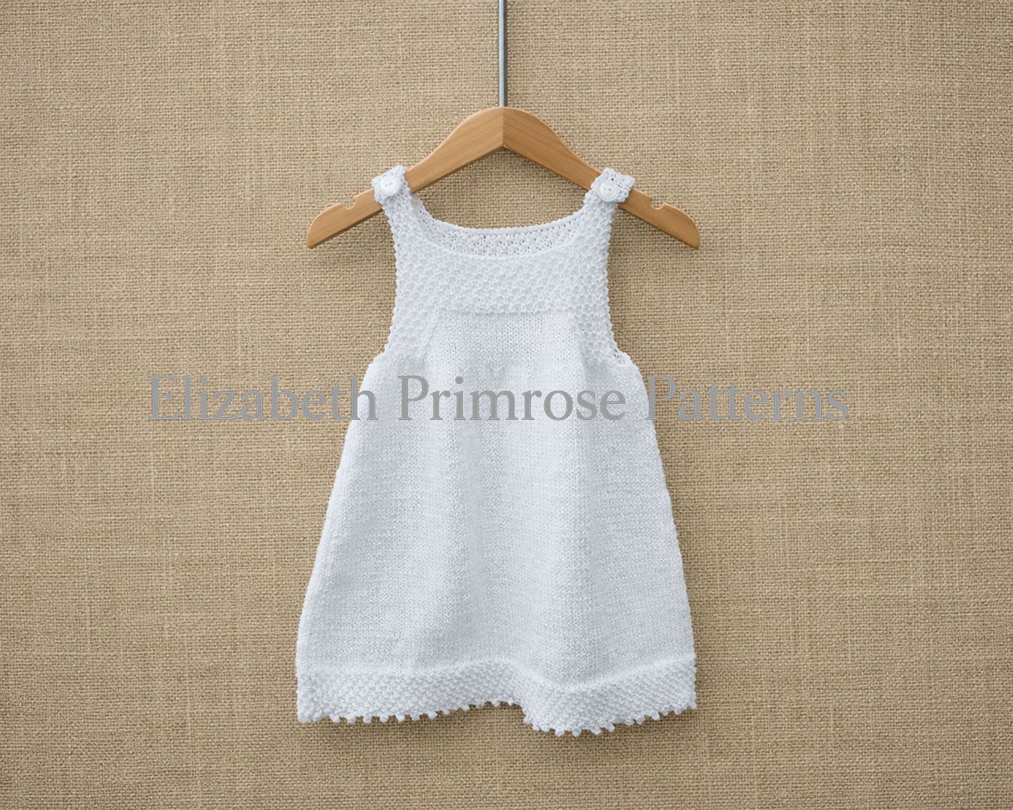 White hand knitted children’s pinafore with textured bodice, buttoned straps and smooth skirt, hanging on a wooden hanger against a soft fabric backdrop.