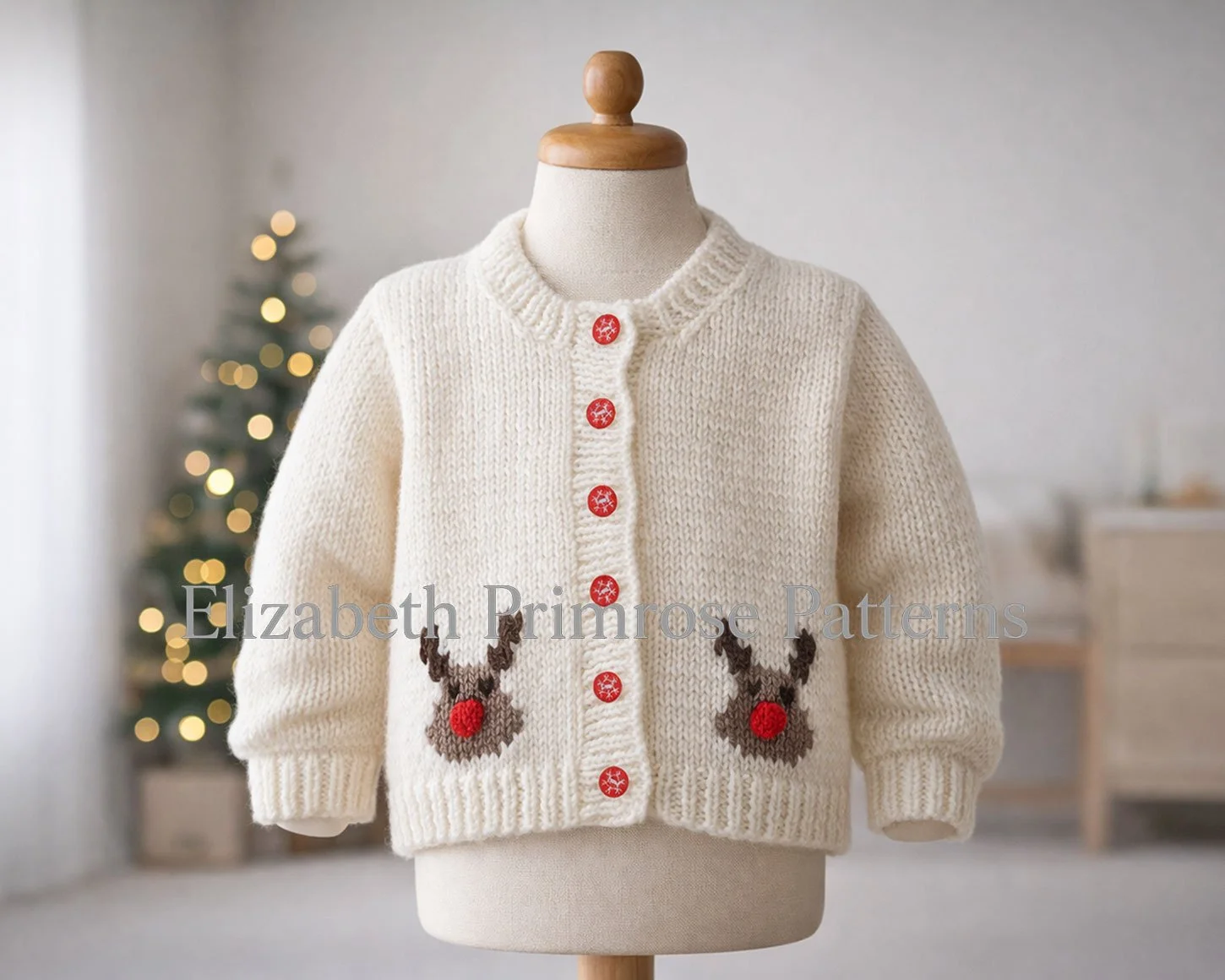 Festive cream children’s cardigan with two Rudolph motifs and red snowflake buttons, displayed on a mannequin against a blurred Christmas tree background.