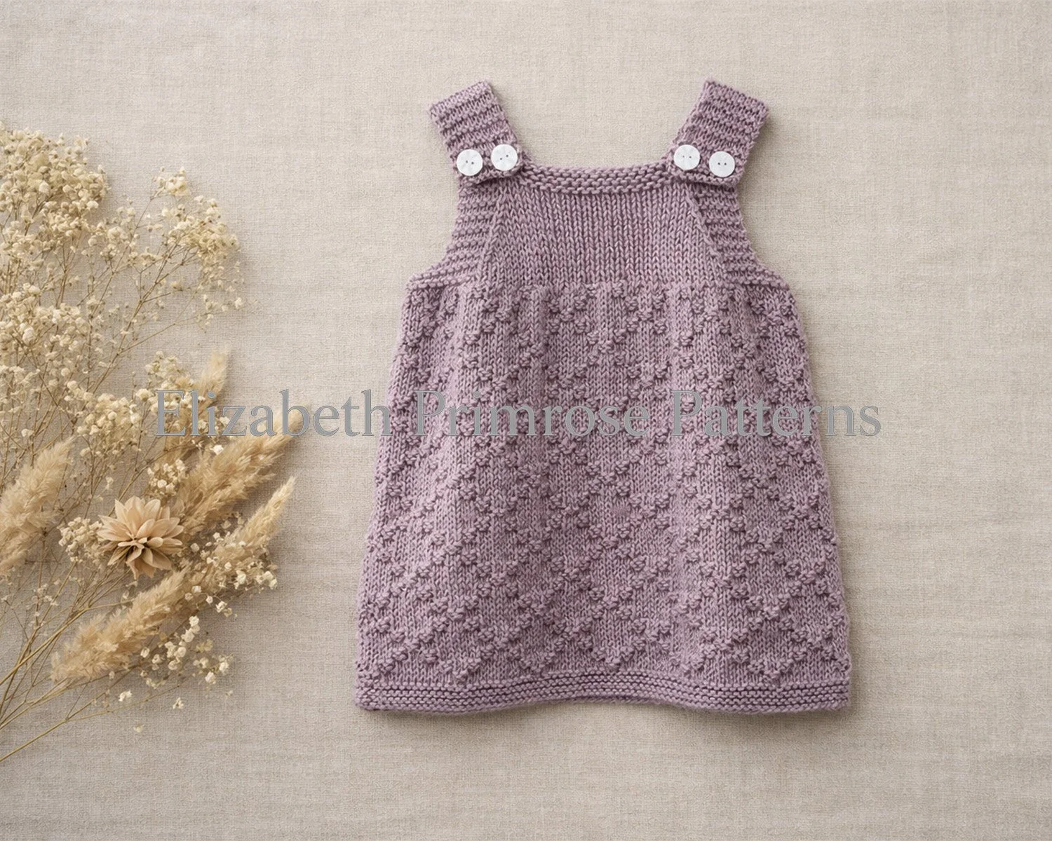 Lavender knitted pinafore dress with diamond texture laid flat on beige linen with soft dried florals beside it. Children’s knitting pattern by Elizabeth Primrose Patterns.