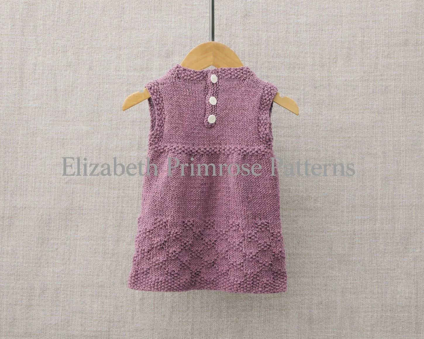 Back view of a hand knitted, sleeveless purple children’s dress on a wooden hanger, showing three back buttons, and textured diamond detailing against beige linen.