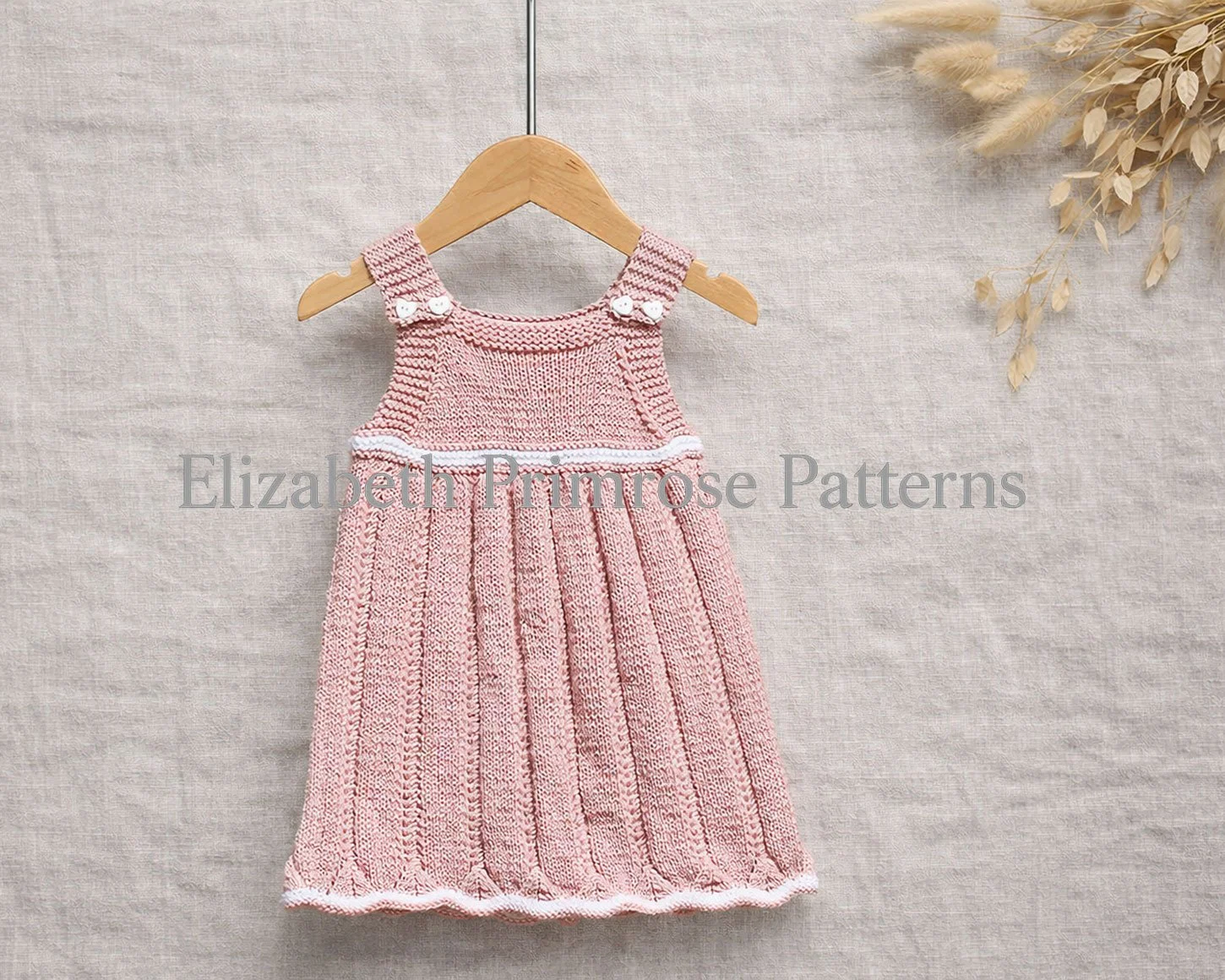 Pink hand knitted baby dress on a wooden hanger against a linen backdrop, showing crisp stitch detail and wavy hem with dried florals beside it.