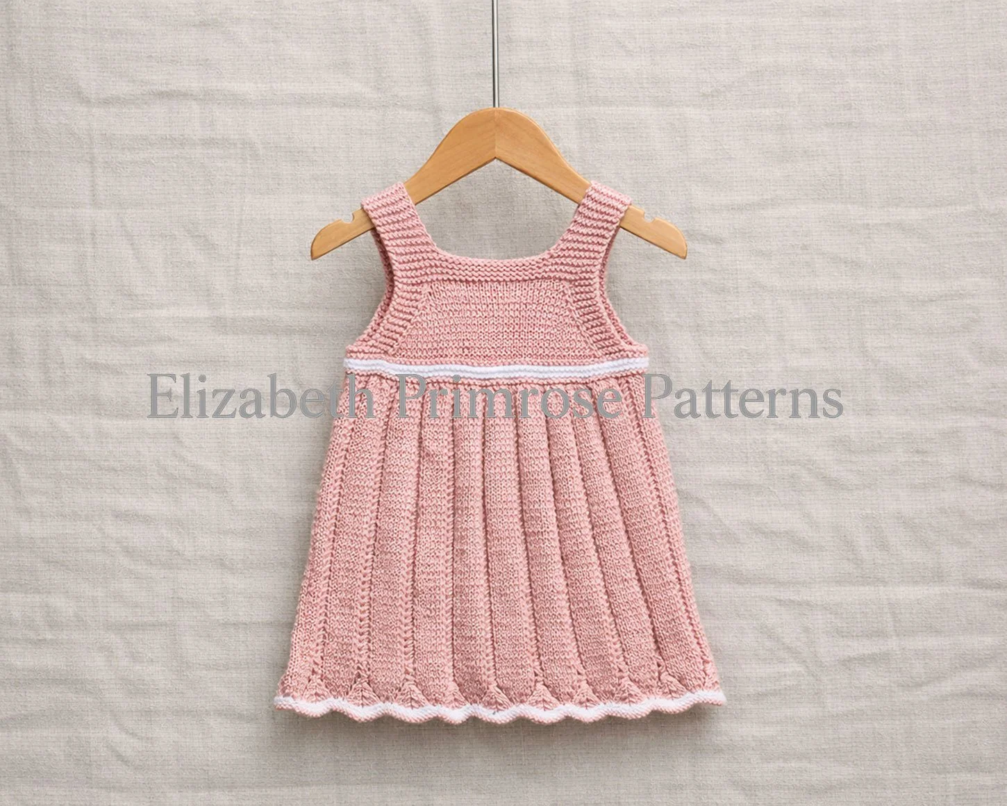 Back view of pink hand knitted children’s dress on a wooden hanger against a linen backdrop.
