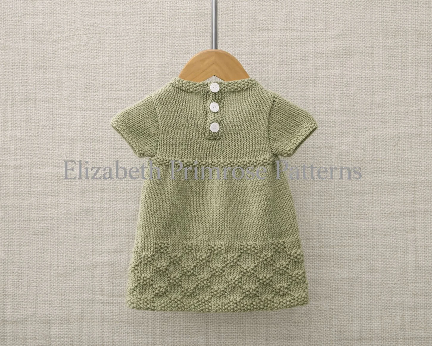 Back view of a hand knitted green children’s dress on a wooden hanger, showing short sleeves, three back buttons, and textured diamond detailing against beige linen.